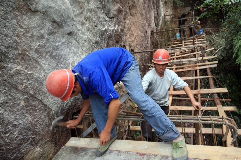 一个正在建设中的悬崖玻璃栈道,农民工在一米多宽的施工面上工作,一边